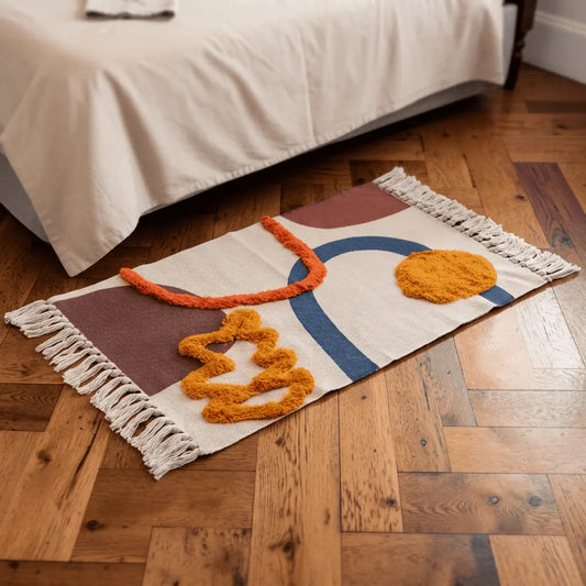 Tapis bohème TERRA écru aux formes abstraites colorées, placé au pied d’un lit sur un parquet chaleureux.