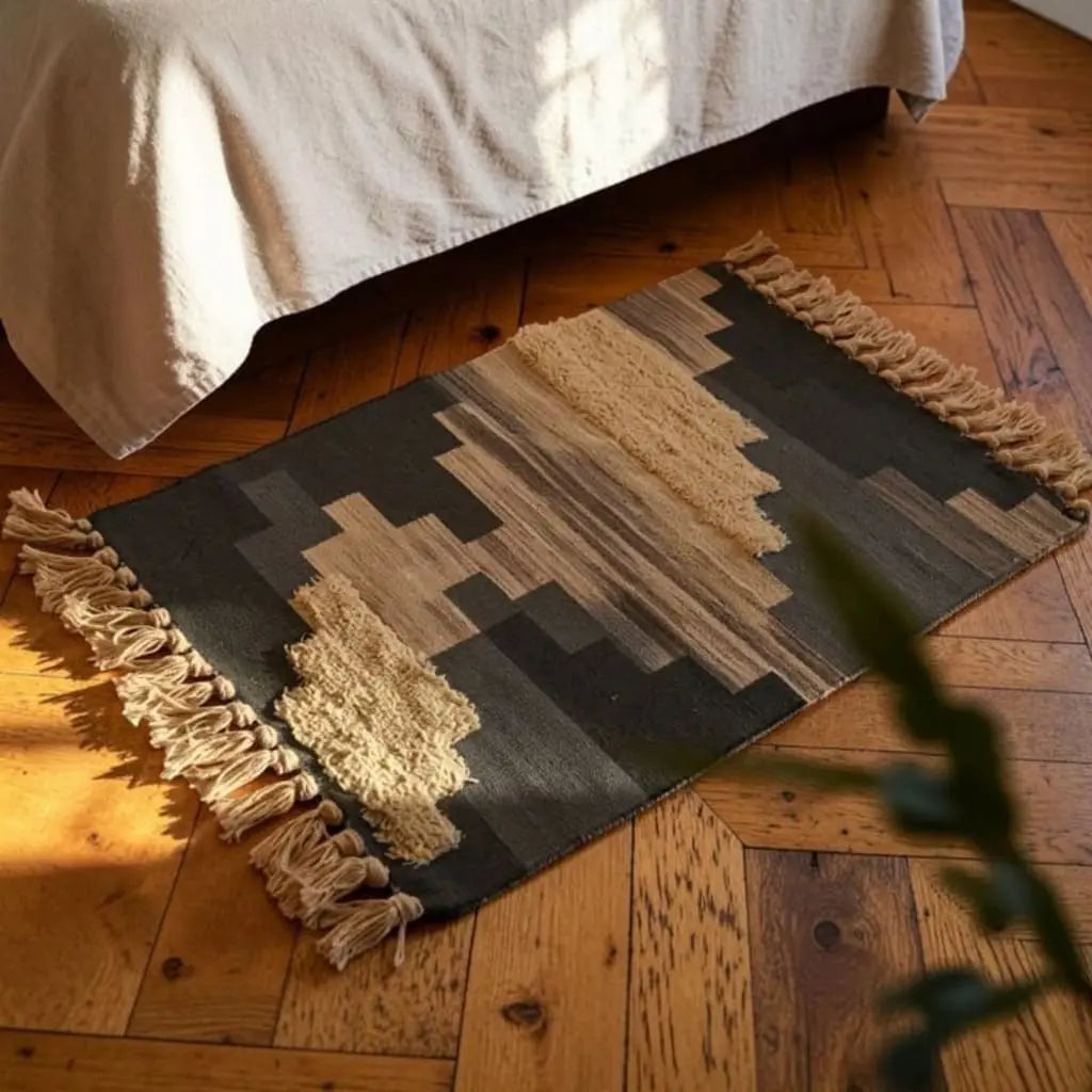Tapis bohème AMAYNO noir et beige à motifs géométriques, placé au pied d’un lit sur un parquet en bois.