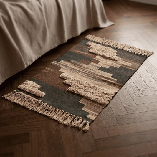 Tapis bohème SIROCCO marron et beige placé au pied d’un lit, sur un parquet à chevrons.