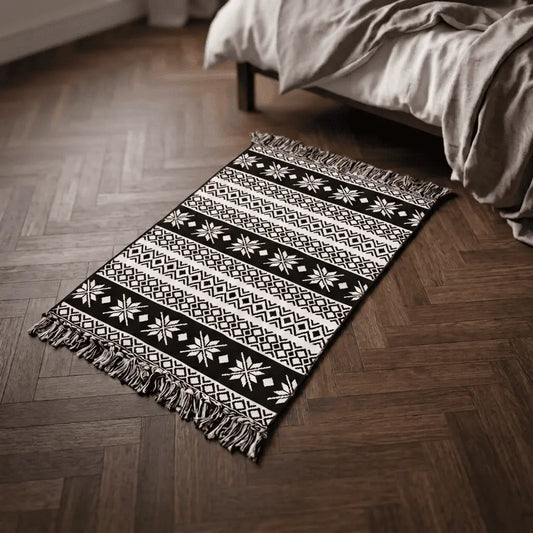 Tapis bohème KENZA noir et blanc placé au pied d’un lit, sur un parquet en bois foncé.